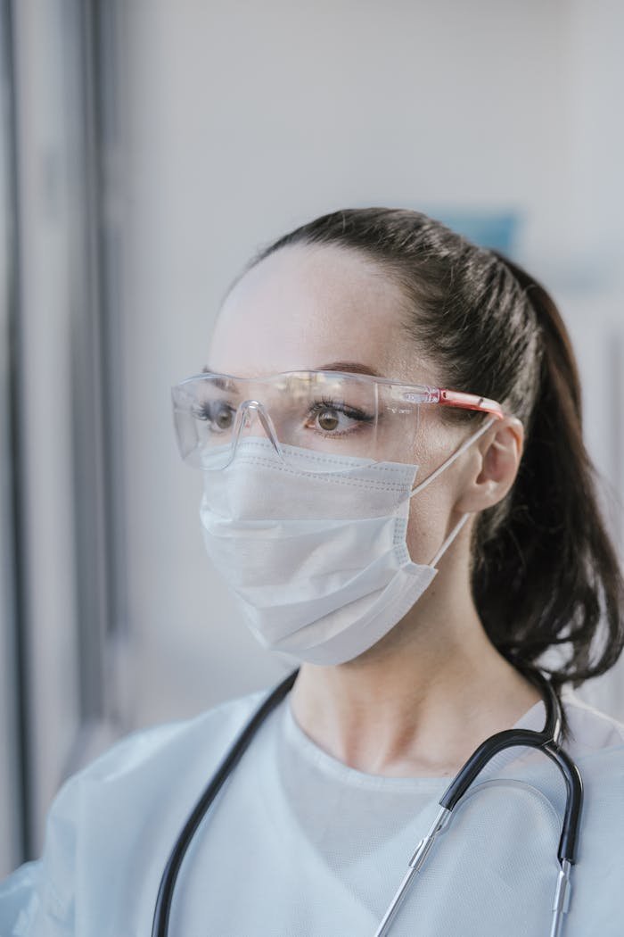 Caucasian female healthcare professional in PPE with a stethoscope looking focused in a clinical environment.